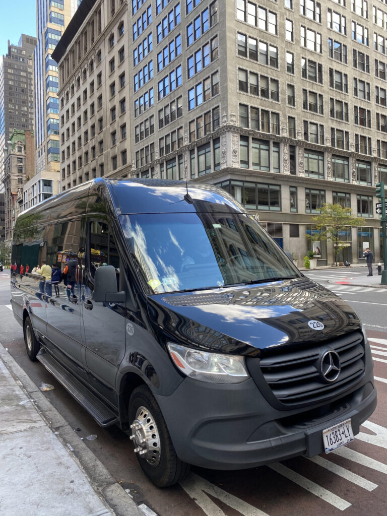 Black Mercedes-Benz van parked at a city street curb, with tall office buildings reflected on the windshield.