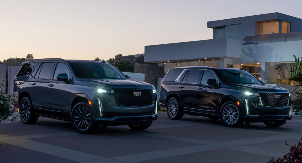 Two Cadillac SUVs parked in a modern driveway at dusk in front of a contemporary house with glass walls and plants nearby.