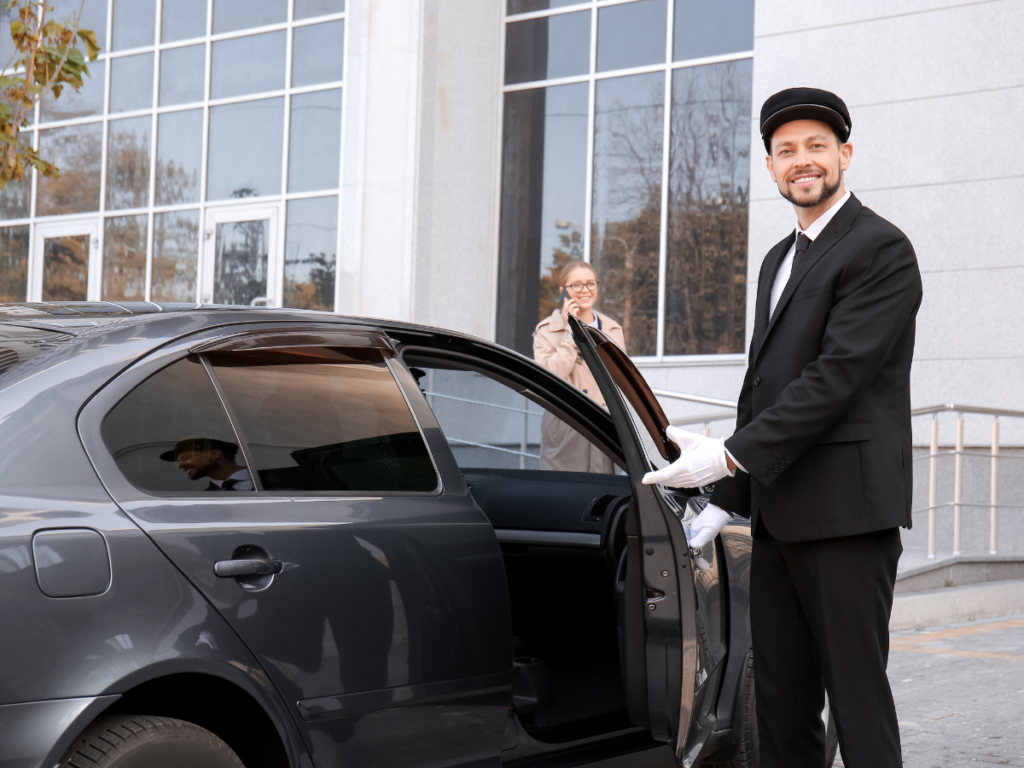 Chauffeur in a suit and cap opens the black car door while a woman on the phone stands near a modern glass-fronted building.