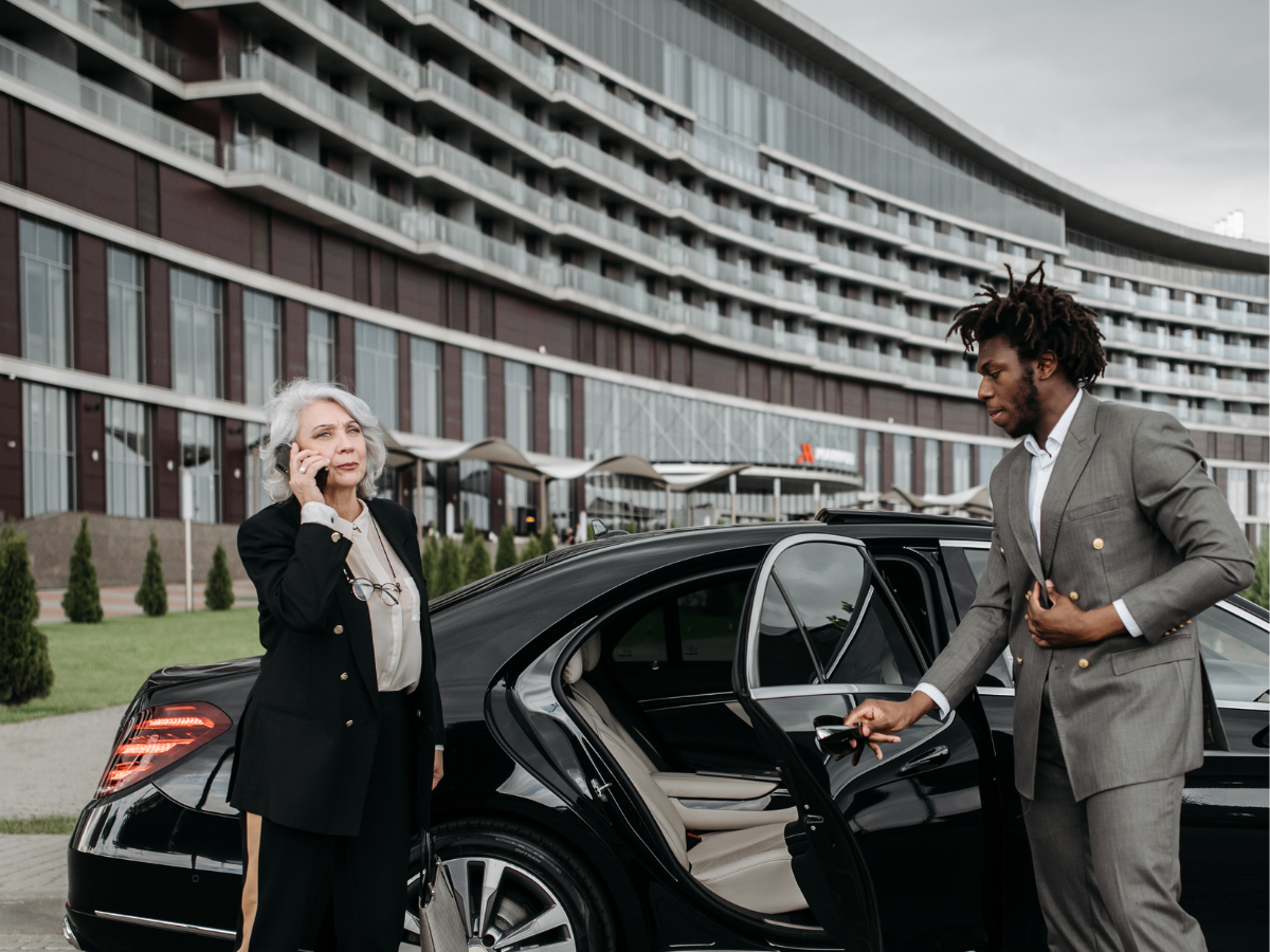 Older woman on a cell phone standing by a black luxury car as a suited chauffeur holds sunglasses at the open rear door, with a curved modern building in the background.