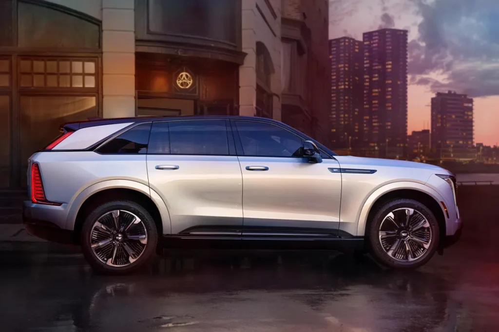 Side view of a silver luxury SUV parked on a city street, with tall buildings at dusk and wet pavement reflecting streetlights.