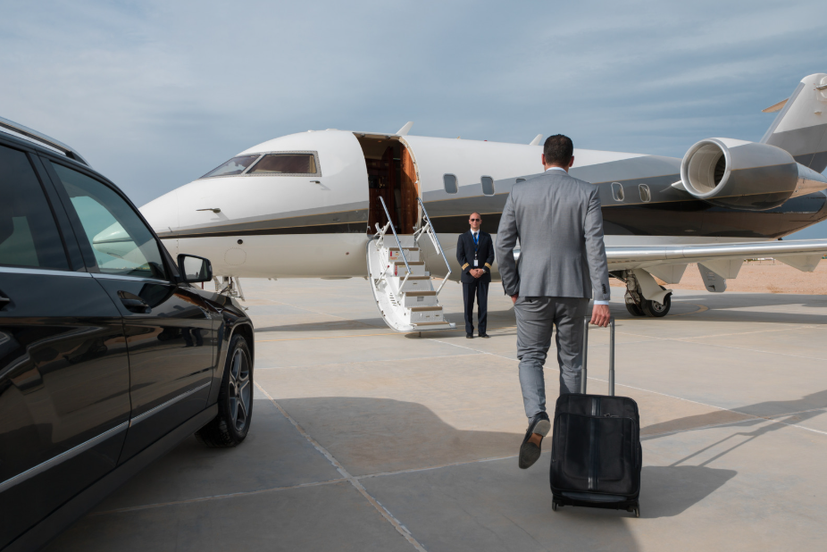 Business traveler in a gray suit walks toward a private jet with a rolling suitcase on a sunlit tarmac, a black car parked nearby.