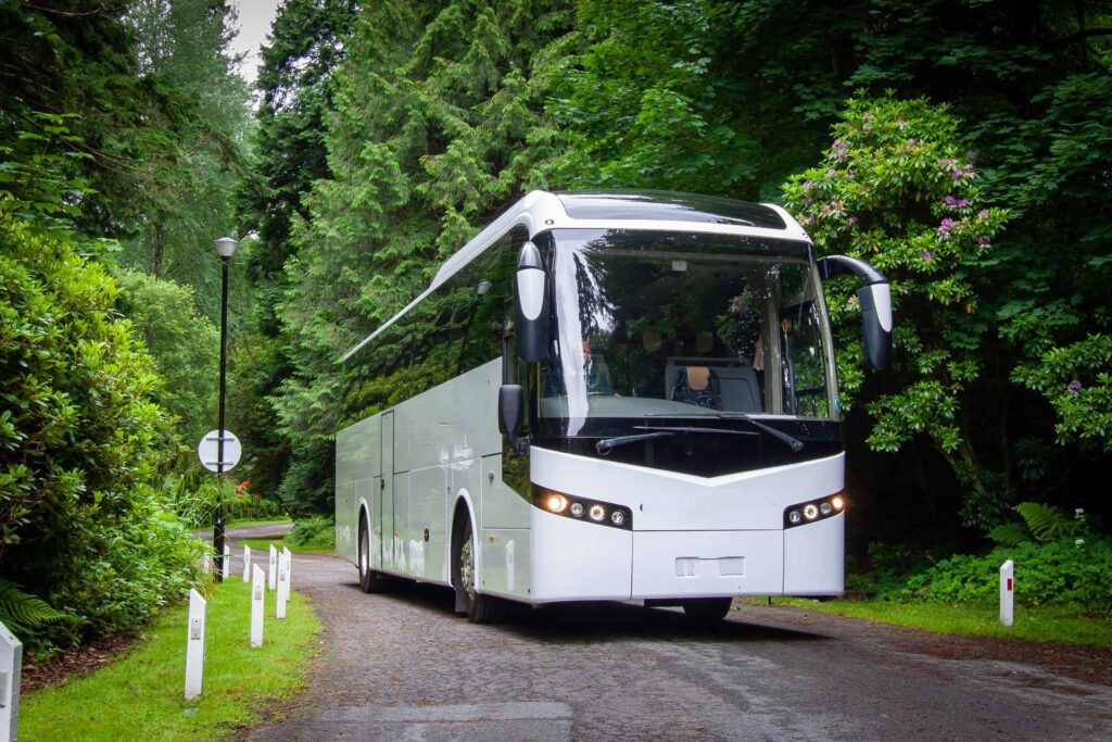 White tour bus driving along a narrow, tree-lined road with dense green foliage on both sides.