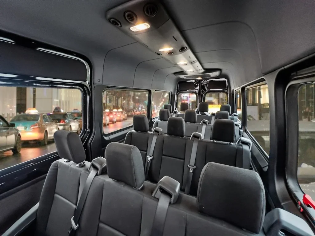 Interior of a multi-row passenger van with grey seats and seat belts, looking toward the back as city street lights appear outside the windows.