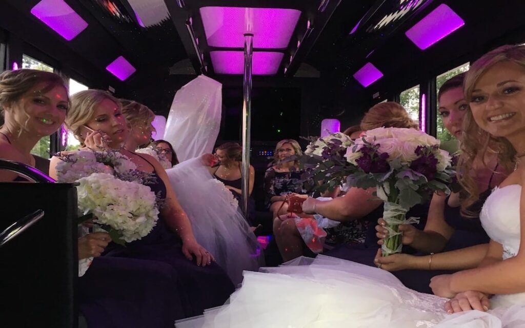 Bride in white dress seated on a party bus with bridesmaids in purple gowns holding bouquets under purple lights.