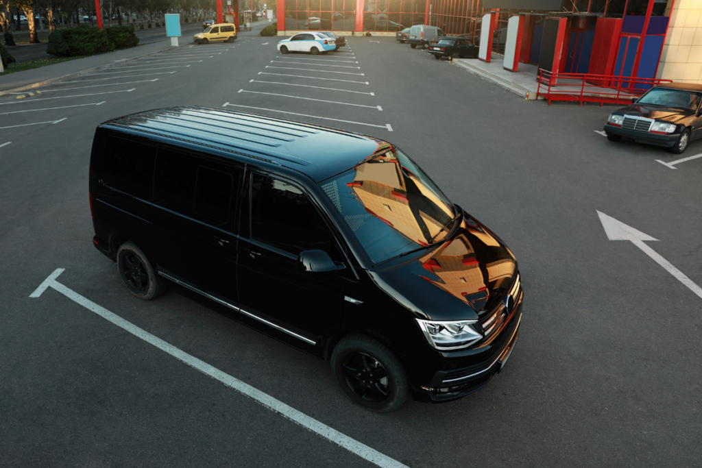 Top-down view of a glossy black van parked diagonally in a mostly empty parking lot outside a commercial building, reflected in its hood.