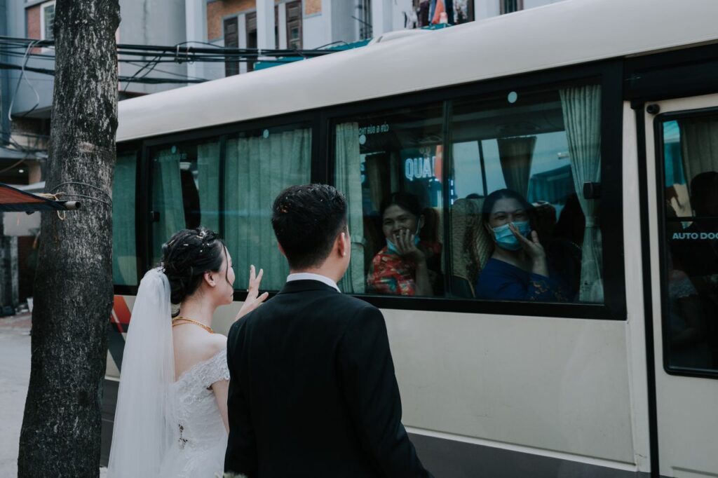 Young bride and groom saying goodbye and waving hands to guests in masks sitting in bus