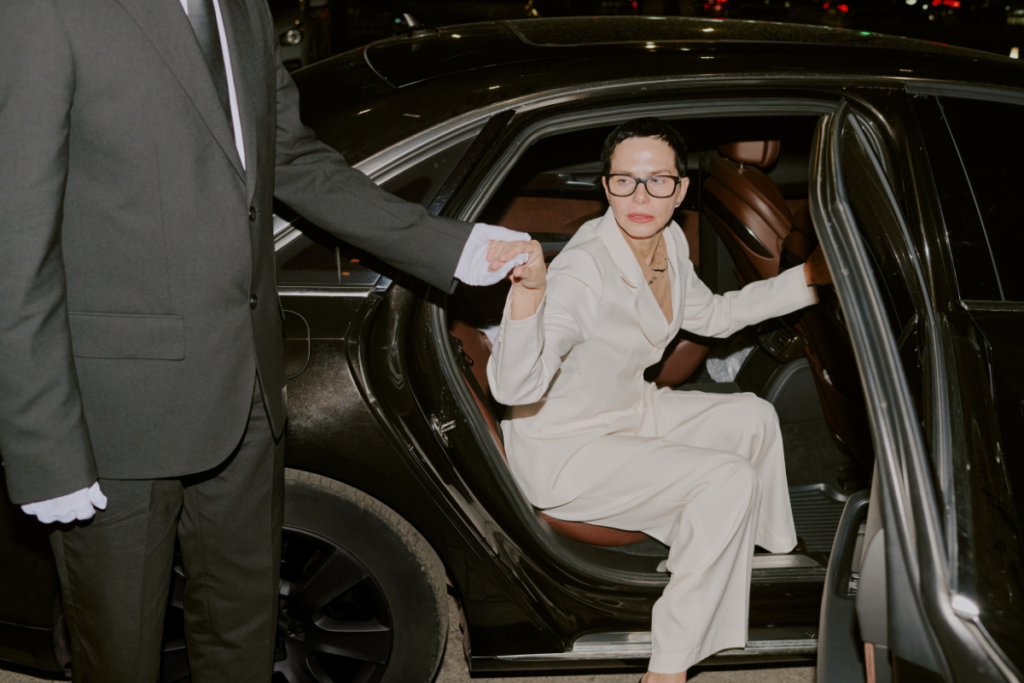 Woman in a white suit stepping out of a black car, guided by a chauffeur in a suit with white gloves.